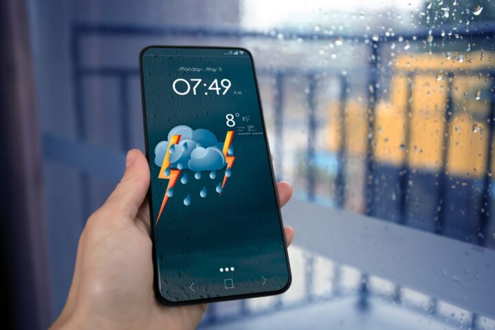 Woman checking weather using app on smartphone near wet window, closeup.
