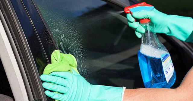 Car owner spraying a glass cleaner on the driver side window with a micro fibre cloth ready to wipe.